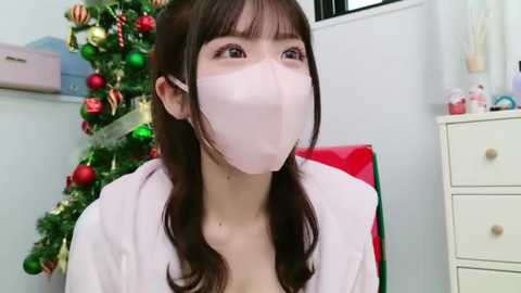 Media: Video of an East Asian woman with long dark hair, wearing a white mask and robe, standing in a festive room with a decorated Christmas tree and red chair.