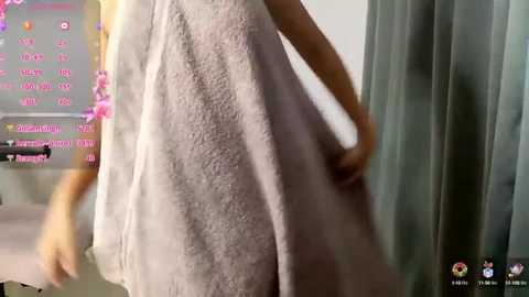 Media: Video of a woman wearing a light gray, textured towel, holding it with both hands. Background includes a digital calendar with pink text, and a gray curtain.