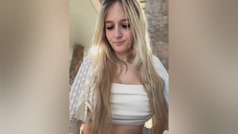 Media: Video of a young, fair-skinned woman with long, straight blonde hair, wearing a white crop top and open-knit cardigan, standing indoors against a brick wall.