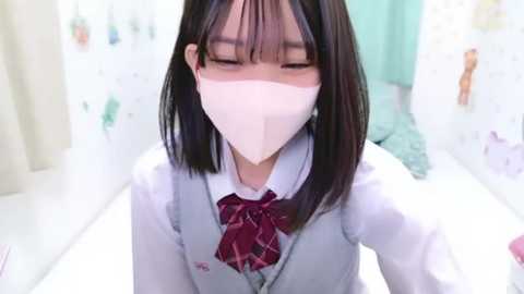 Media: A video of a young Asian girl with straight black hair and bangs, wearing a white school uniform with a red plaid bow tie, a white face mask, and a light blue cardigan. She is indoors, with blurred pastel-colored walls and a green towel in the background.