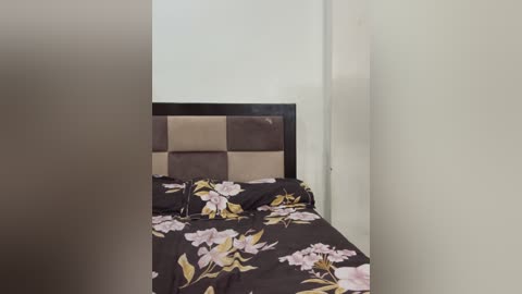 Media: Video of a small, simple bedroom with a dark floral bedspread, a padded beige headboard, and plain white walls. The room appears empty and minimalistic.