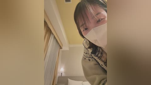 Media: A video of a young Asian woman with long dark hair, wearing a white mask, looking out from a dimly-lit, beige-walled hallway.