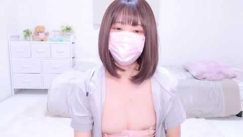 Media: Video of an East Asian woman with short, straight brown hair, wearing a white mask, light grey shirt, and pink bra, in a minimalistic, white bedroom with a bed and dresser.