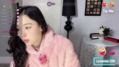 Media: Video of a young Asian woman with long black hair, wearing a pink fuzzy robe, in a cozy bedroom with white walls, a black lamp, and a white table.