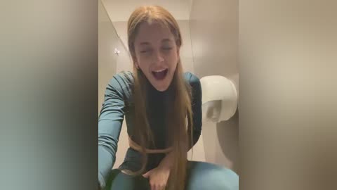 Media: Video of a young, Caucasian woman with long, straight, light brown hair, wearing a teal sweater and blue pants, laughing while leaning forward in a public restroom with beige walls and a white toilet paper dispenser.