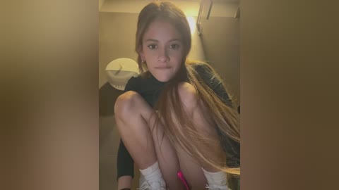 Media: Video of a young, light-skinned woman with long, straight blonde hair, squatting in a dimly lit room. She wears a black sweater and pink panties, with white socks. Background features a beige wall, a white lamp, and a small, indistinct object.