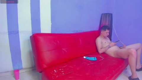 Media: A video of a naked, fair-skinned, slender man with short dark hair, sitting on a red couch, holding a smartphone, in a blue and white striped room.