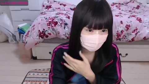 Media: Video of a young woman with black hair, wearing a face mask, black jacket, and touching her hair, seated on a colorful rug in a bedroom with a bed covered in floral bedding.