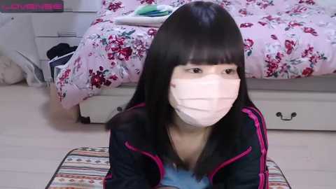 Media: A video of an Asian woman with long black hair, wearing a pink face mask, seated on a patterned rug in a pink floral bedroom.