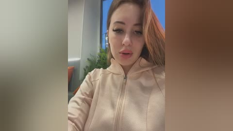 Media: A video of a young, fair-skinned woman with straight brown hair, wearing a beige zip-up hoodie, taking a selfie indoors near a window with green plants outside.