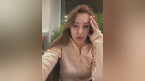 Media: Video of a Caucasian woman with long, light brown hair, wearing a beige zip-up jacket, looking tired, with a hand on her forehead, blurred background.