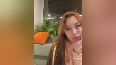 Media: A close-up video of a young woman with long, straight, light brown hair, wearing a light pink top, resting her chin on her hand, in a modern interior with a potted plant and orange cushion.