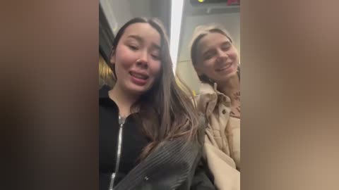 Media: Video of two young women, one with long dark hair and a black jacket, the other with blonde hair and a beige jacket, smiling, in an elevator.