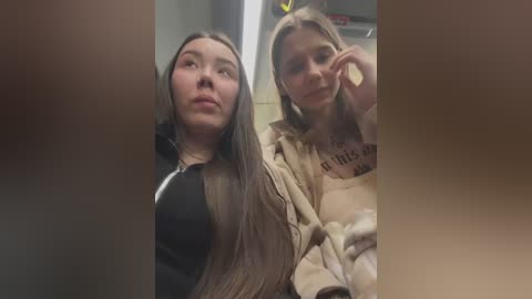 Media: Video of two young women in a dimly lit room, one with long dark hair, black top, and pale skin, the other with blonde hair, beige top, and lighter skin.