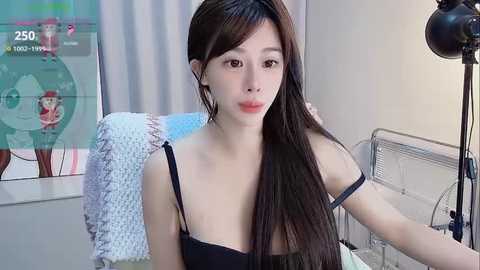 Media: Video of a young Asian woman with long, straight brown hair, wearing a black spaghetti-strap top, sitting in a modern, minimalistic room with a white chair and a camera on a tripod.