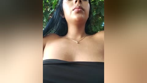 Media: A video of a woman with medium skin tone and long black hair, wearing a strapless black top, standing outdoors against a lush green leafy background. She has a slender physique and a neutral facial expression.