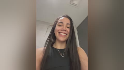 Media: Video of a smiling Latina woman with long black hair, wearing a black sleeveless top, standing in a modern indoor setting with neutral-toned walls and a ceiling vent.