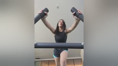 Media: Video of a fit woman with long dark hair, wearing a black tank top and blue shorts, lifting a black barbell in a home gym with a wooden floor and beige walls.