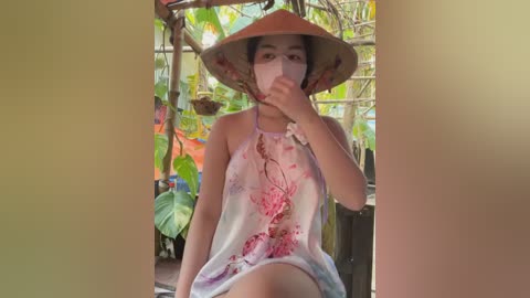 Media: Video of a young Asian woman with medium skin tone, wearing a pink floral-patterned halter top and a conical hat, sitting outdoors in a garden with green foliage and hanging plants.