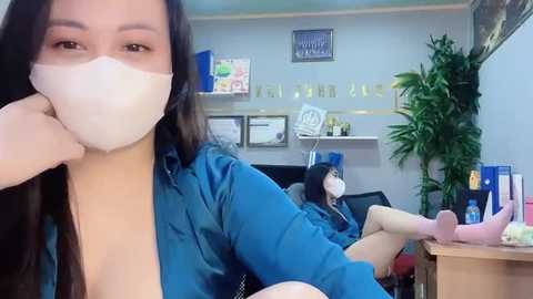 Media: Video of an Asian woman with long black hair wearing a blue jacket, mask, and glasses, taking a selfie in a doctor's office with a patient in the background.