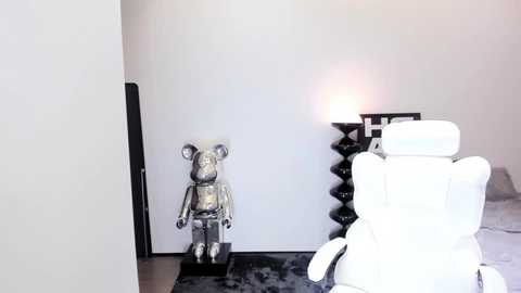 Media: A video of a minimalist room with a white plush chair facing a wall adorned with a metallic, cartoonish robot figure and a black, geometric lamp.