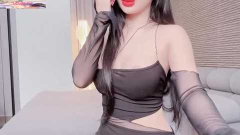 Media: Video of a woman with long black hair, wearing a black sheer top, elbow-length gloves, and red lipstick, standing in a modern bedroom with a grey bed and beige wallpaper.