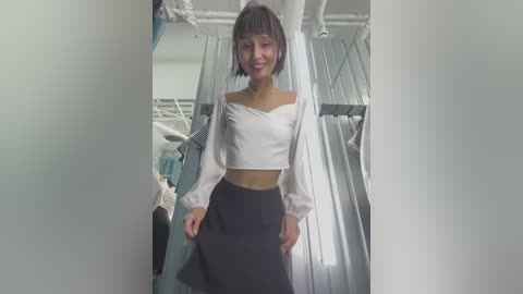 Media: A video of a young woman with short, straight hair and fair skin, wearing a white off-the-shoulder crop top and high-waisted black skirt, standing in a brightly lit, modern clothing store with hanging garments in the background.