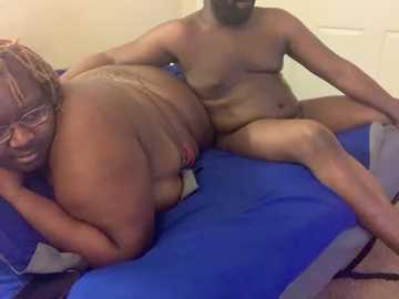 Media: Video of a nude, dark-skinned woman with glasses and a plump body, lying on a blue bed. A bearded, muscular dark-skinned man is partially visible, sitting behind her.