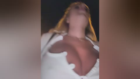 Media: Video of a blonde woman with large breasts, wearing a low-cut white top, from a low angle, blurred background.