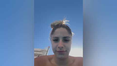 Media: Video of a woman with fair skin and blonde hair tied in a messy bun, wearing a pink top, standing in front of a bright blue sky.