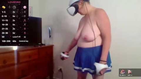 Media: Video of a topless, light-skinned woman wearing a VR headset, blue skirt, and white gloves, holding a VR controller, standing in a sparsely furnished room with a TV and a wooden dresser.