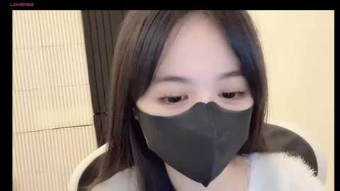 Media: A close-up video of an Asian woman with long, straight black hair, wearing a black face mask and a white fur-trimmed coat, sitting indoors with beige walls and a wooden screen in the background.