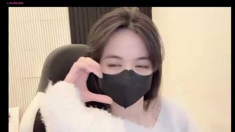 Media: Video of an Asian woman with long brown hair, wearing a black face mask and white fluffy robe, speaking on a black phone, indoors with beige walls and a vertical wooden screen.