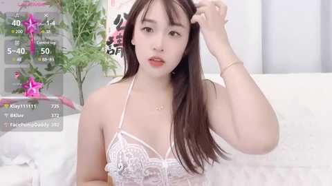 Media: Video of an East Asian woman with long black hair, wearing white lace lingerie, adjusting her hair in a modern, minimalist room.