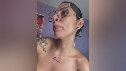 Media: Video of a topless woman with medium skin tone, brown hair tied back, wearing glasses, and black hoop earrings, with tattoos on her left shoulder and chest. Background features purple walls and framed artwork.