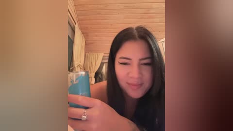 Media: Video of a young Asian woman with long black hair, wearing a black top, smiling while holding a blue drink in a clear glass. The background features wooden ceiling panels and beige curtains.