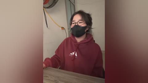 Media: Video of a woman in a red hoodie, black mask, and glasses, standing in a dimly lit, beige-walled room with a wooden table, a ceiling fan, and a yellow cord.