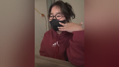 Media: Video of an East Asian woman with glasses and a black face mask, wearing a red hoodie, holding a phone in a dimly lit room.