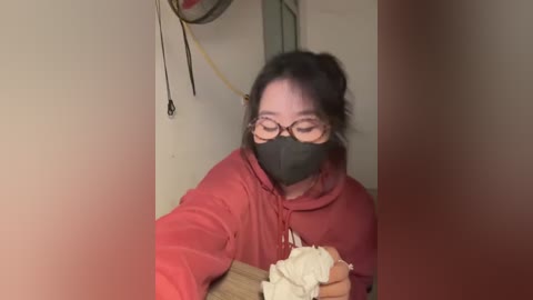 Media: Video of an Asian woman with glasses and a black mask, wearing a red hoodie, eating white noodles indoors. Background shows a wall and a hanging object.