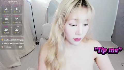 Media: A video of a young, fair-skinned, blonde woman with long hair, wearing only a pink lipstick, taken in a minimalist, white room. A virtual tip jar with \"$tip me\" text overlays the image.