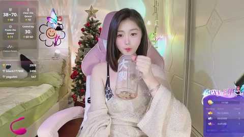 Media: Video of an East Asian woman with straight black hair, wearing a beige robe, drinking from a glass jar, in a cozy room with a decorated Christmas tree.