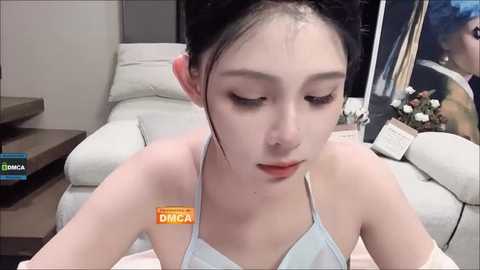 Video of a young Asian woman with pale skin and dark hair, wearing a light blue halter top, sitting on a white couch in a modern living room.