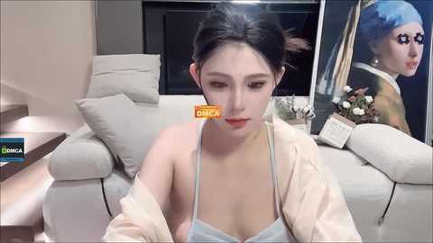 Media: Video of a young East Asian woman with pale skin, dark hair, and minimal makeup, wearing a light blue tank top and beige jacket, sitting on a gray couch. Background features a framed painting and a bookshelf.