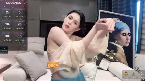 Media: Video of an Asian woman in a beige top, adjusting a blue-haired mannequin's hair, in a modern living room with a TV showing a weather forecast.