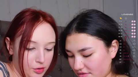 Media: Video of two young women with fair skin, one with red hair, one with black hair, both with closed eyes, resting on a grey cushioned surface.