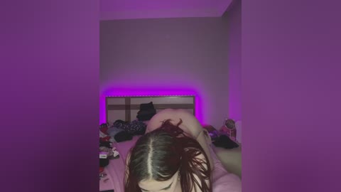 Video of a woman with long brown hair lying face down on a bed in a dimly lit room, bathed in purple lighting. The room is cluttered with clothes and personal items.