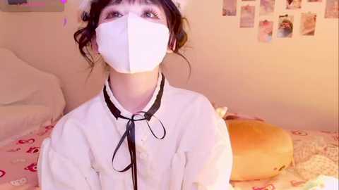 A video of a young woman with pale skin and dark hair wearing a white maid costume, including a face mask, in a cozy, softly lit bedroom.