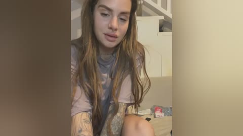 Media: A young woman with long brown hair, light skin, and a slight smile sits on a bed, wearing a gray shirt and revealing tattoos on her arms. The background shows a white staircase and a cluttered nightstand.