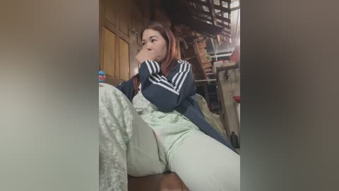 Video of a young woman with long red hair, wearing a dark blue Adidas jacket and light green leggings, sitting on a bed in a cluttered room with wooden walls.