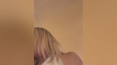 Media: Video of a blonde woman with shoulder-length hair, partially obscured by a beige wall, possibly in a bathroom. She appears to be wearing a white shirt, and the image has a soft focus.